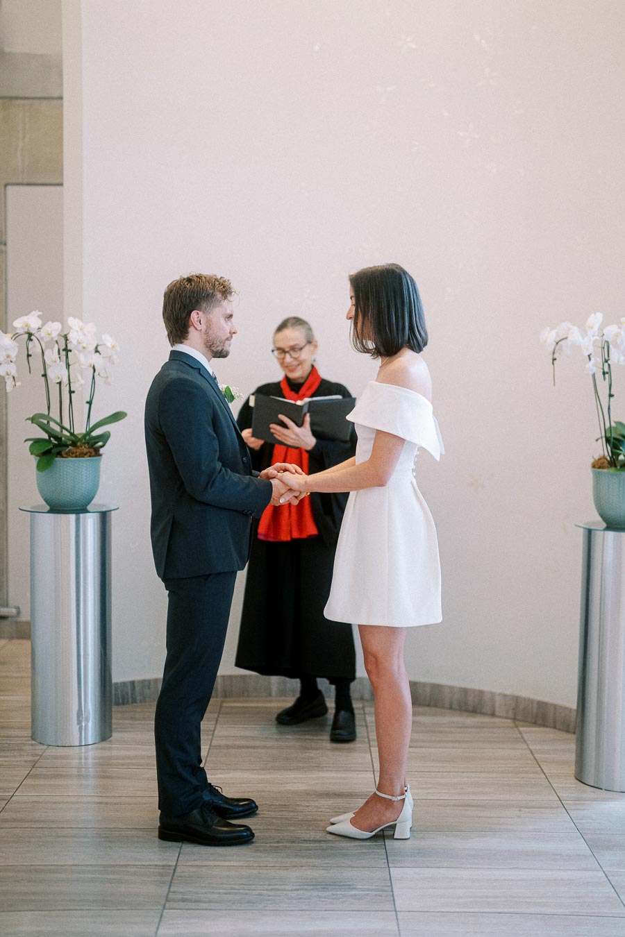 A couple exchanging vows during an indoor wedding ceremony, with a officiant in the background holding a book. The bride is wearing a white dress, and the groom is in a dark suit. Elegant decor includes white flowers on either side.