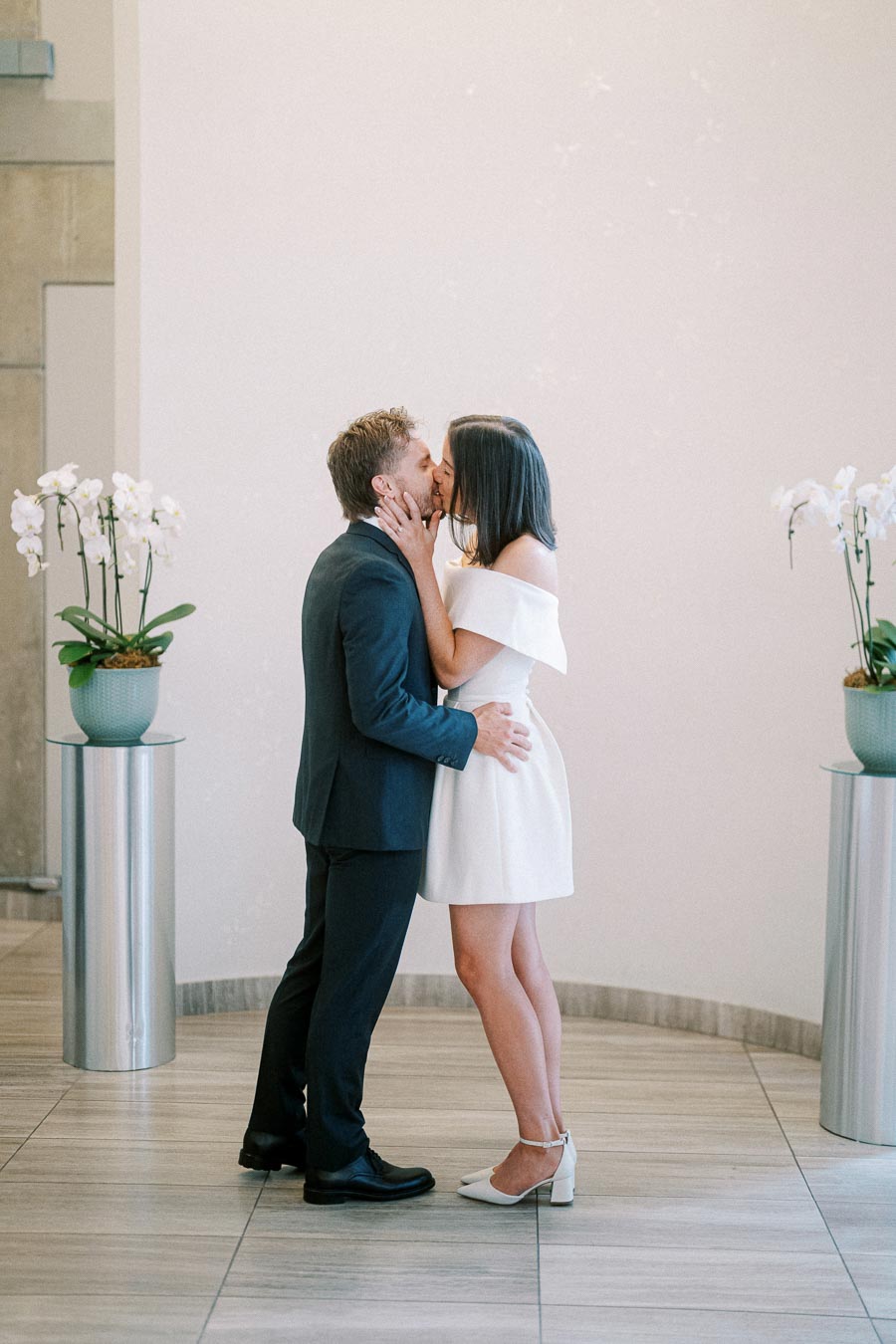 A couple sharing a romantic kiss during their wedding ceremony in an elegant indoor setting with orchid flower arrangements.