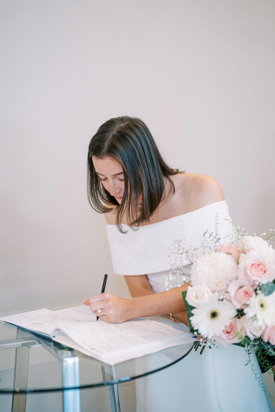 A bride in an elegant white dress signing a marriage certificate, with a bouquet of pink and white flowers on a glass table.