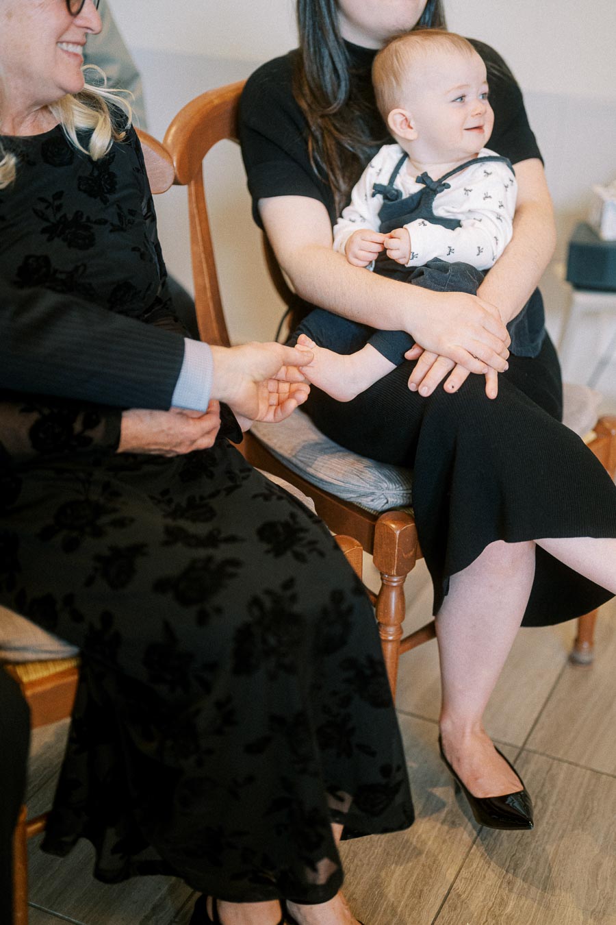 A baby in a patterned white shirt and navy overalls sits on a woman's lap, smiling and looking to the side. Another woman is seated beside them, gently holding the baby’s foot. Both women are dressed in black attire and are seated on wooden chairs.