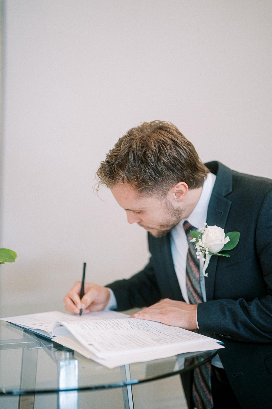 A groom in a suit signing a wedding register at a ceremony, wearing a boutonniere with a white rose, symbolizing marriage and commitment.