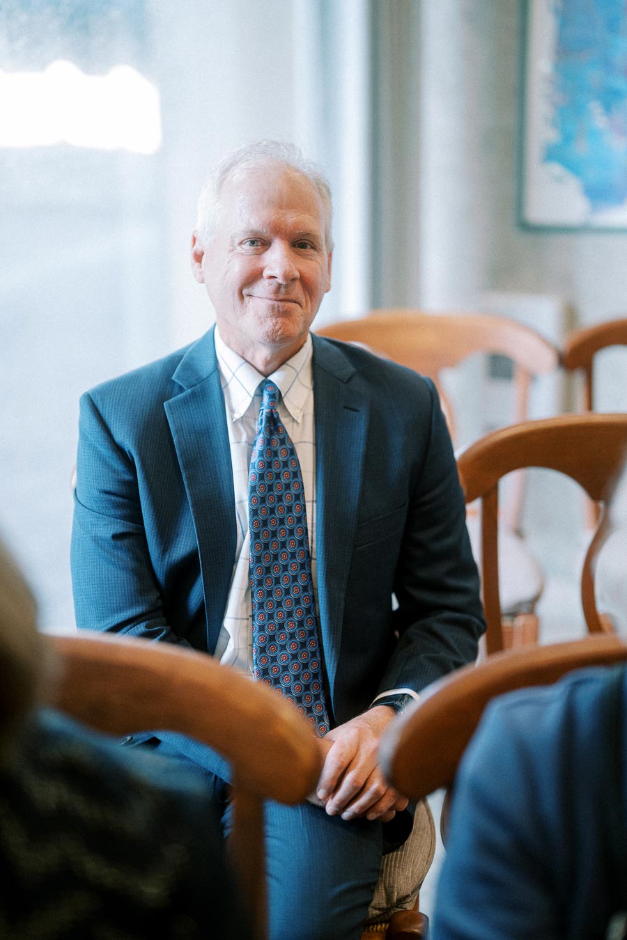 A professional man in a blue suit with a patterned tie sitting on a wooden chair in a bright office setting.
