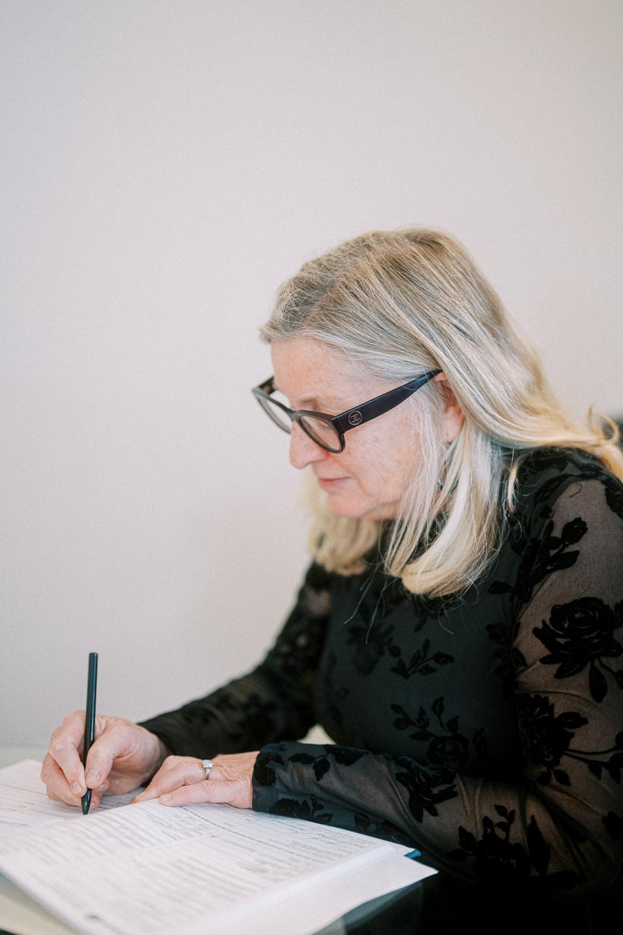 Elderly woman with glasses writing on paper at a desk, wearing a black floral patterned blouse, focused on her task.