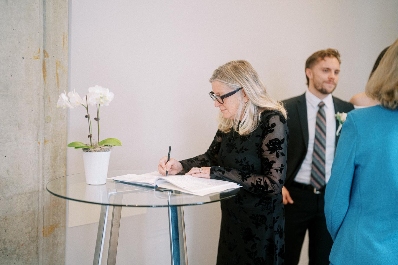 A woman in a black floral dress signs a guest book on a glass table adorned with a white orchid, with people standing nearby in a modern indoor setting.