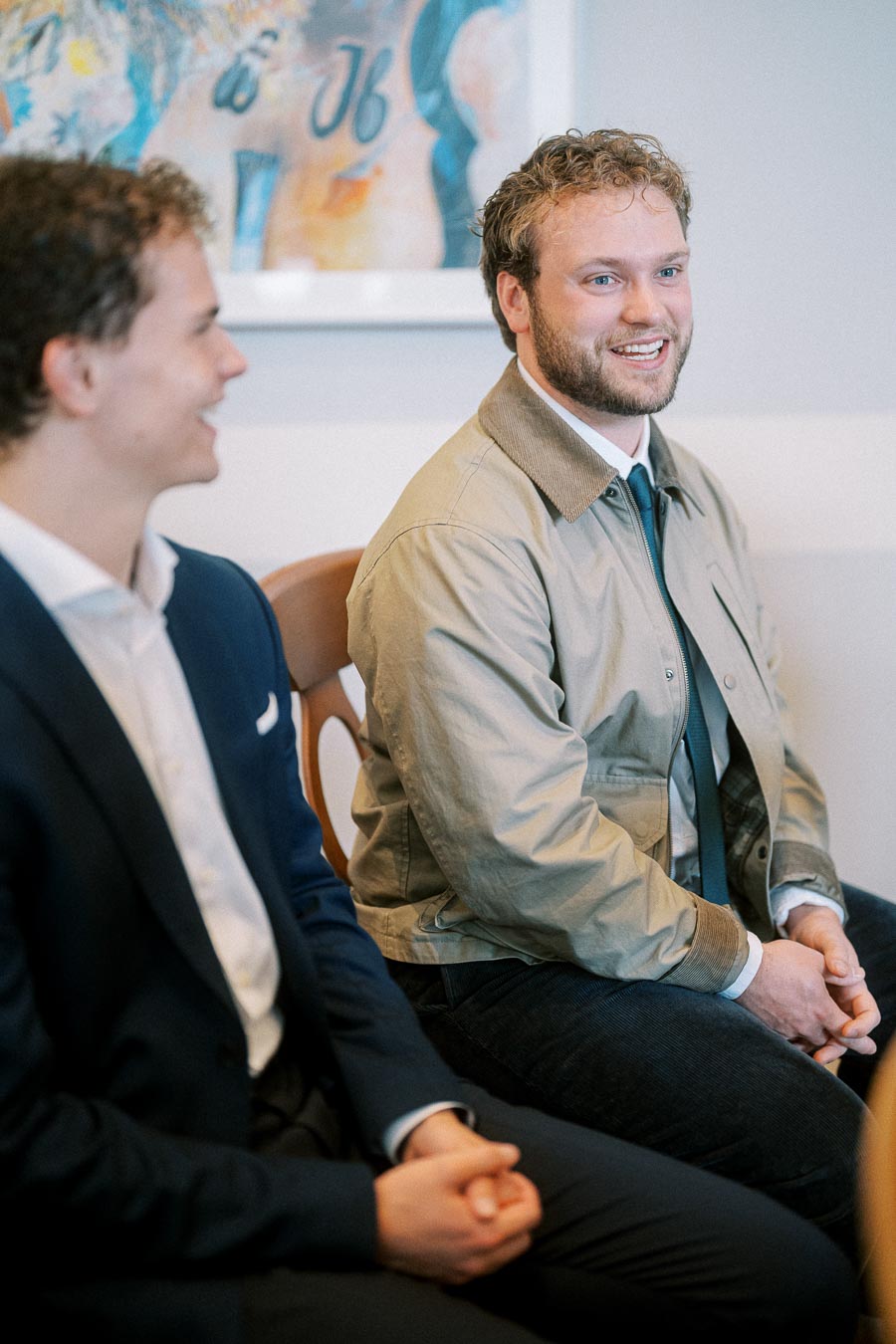 Two men sitting and talking, one wearing a beige jacket and the other in a suit, in a business setting.