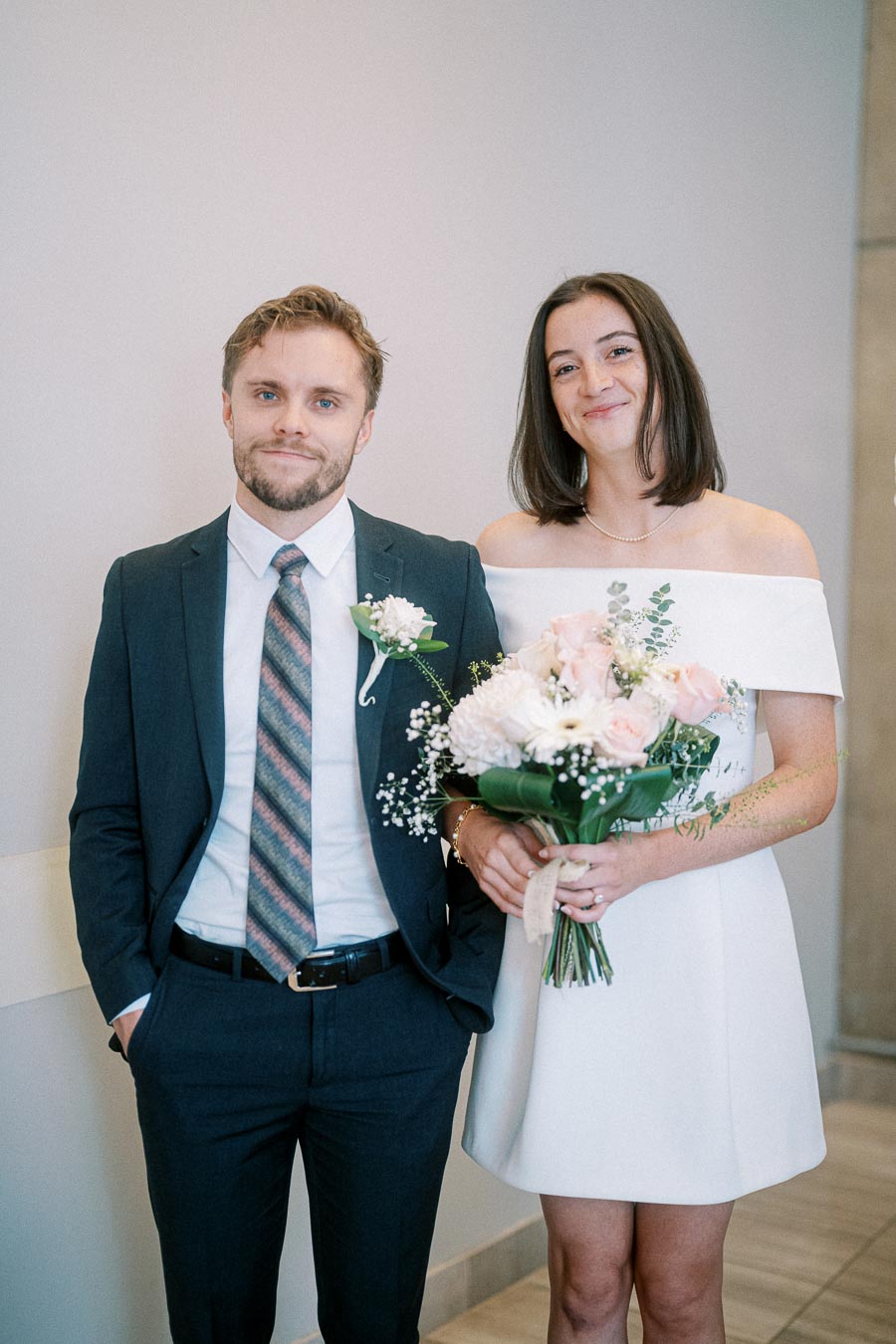 Couple in wedding attire posing indoors, with the woman holding a bouquet of flowers and the man wearing a suit and tie, standing against a neutral background.