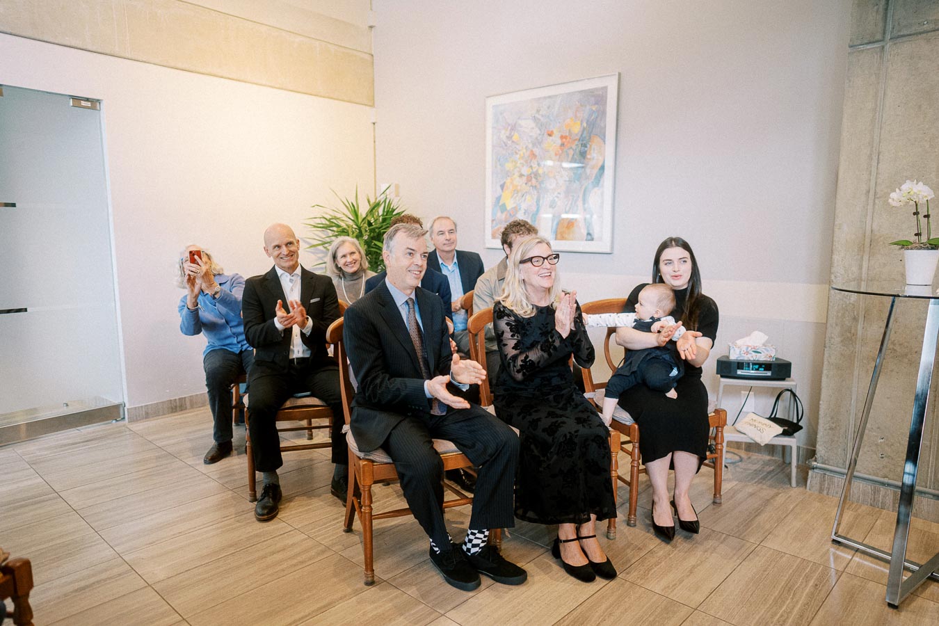 Family gathering in a stylish modern setting with adults and a baby. Attendees, dressed in formal attire, are seated on wooden chairs, smiling and applauding. A mix of generations adds warmth to the intimate event.