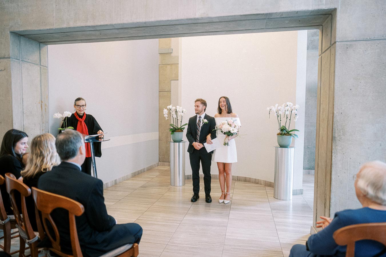 A couple standing together in a modern wedding ceremony setting, with a smiling officiant in a red scarf and guests watching from wooden chairs. The bride holds a bouquet and wears a white dress, while the groom is in a suit, standing in a minimalist room adorned with elegant potted orchids.