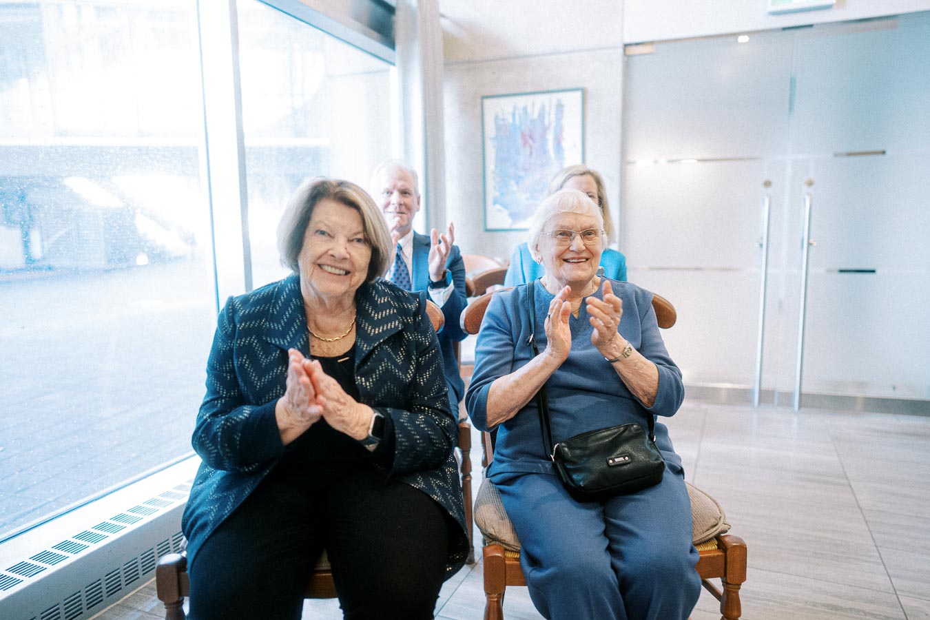 Elderly women and men smiling and clapping while sitting on chairs in a bright room with large windows.