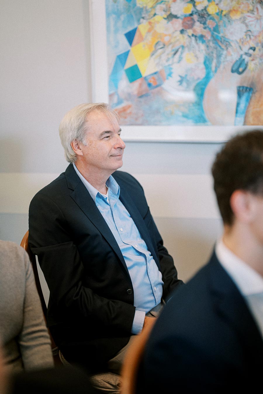 A man in a dark suit sitting attentively at a meeting, with an abstract colorful painting in the background.