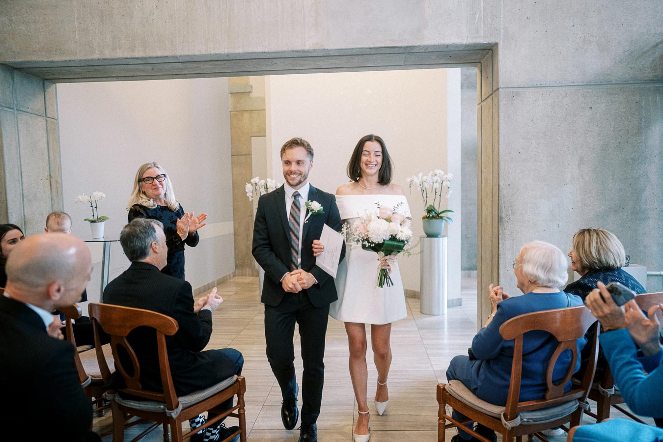 Newlywed couple exiting wedding ceremony, surrounded by applauding guests, in a modern venue with minimalist decor and orchids.