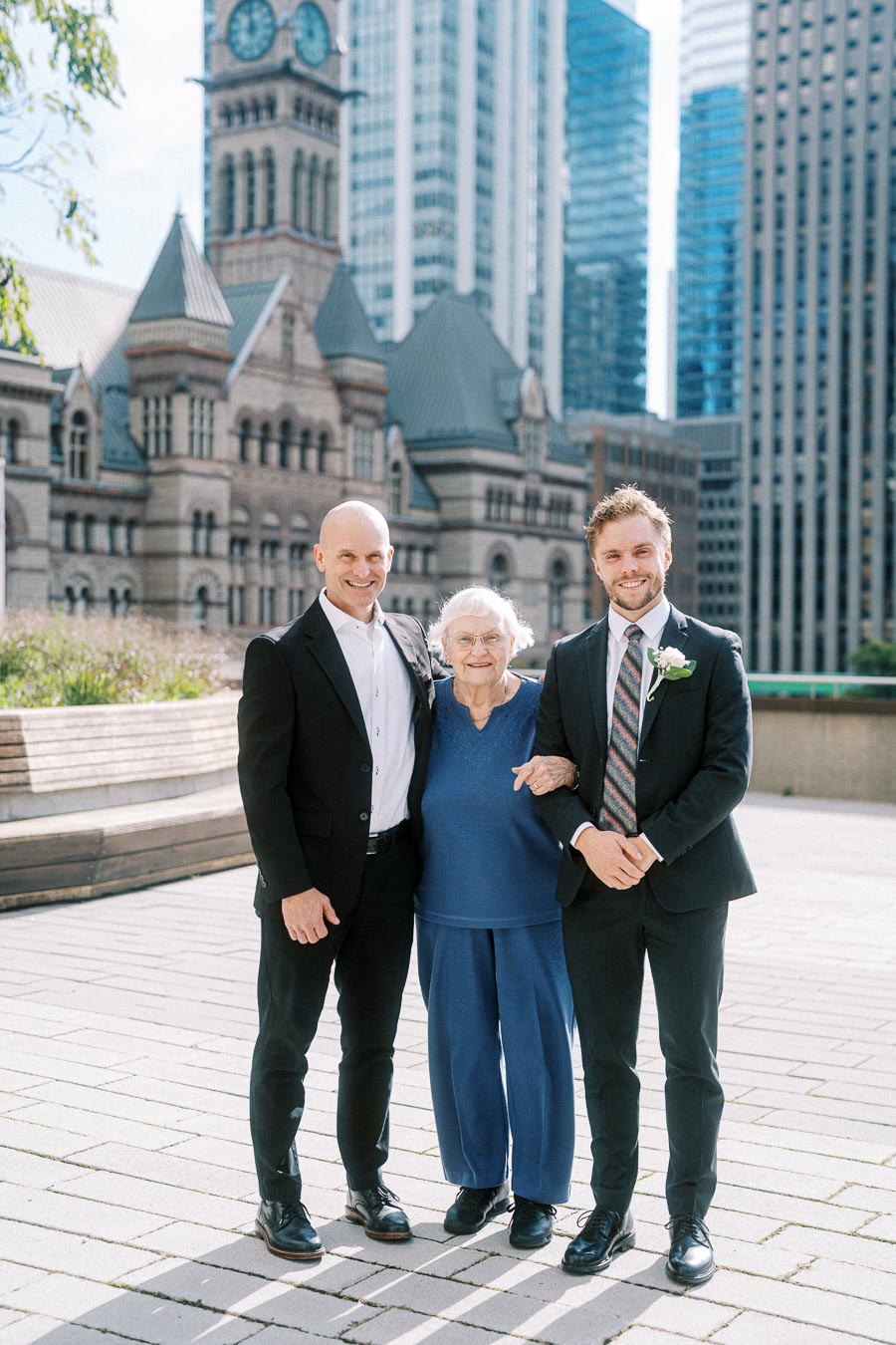 Three people standing together in front of a historic building with a large clock tower, surrounded by modern skyscrapers on a sunny day.