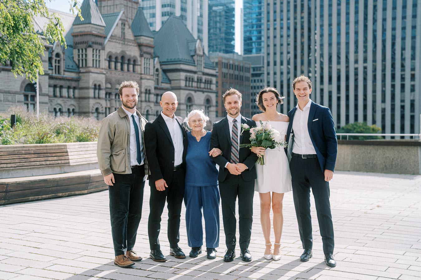 A group of people posing in formal attire at an outdoor event with historic and modern buildings in the background. The woman in a white dress holds a bouquet of flowers, suggesting a wedding celebration.