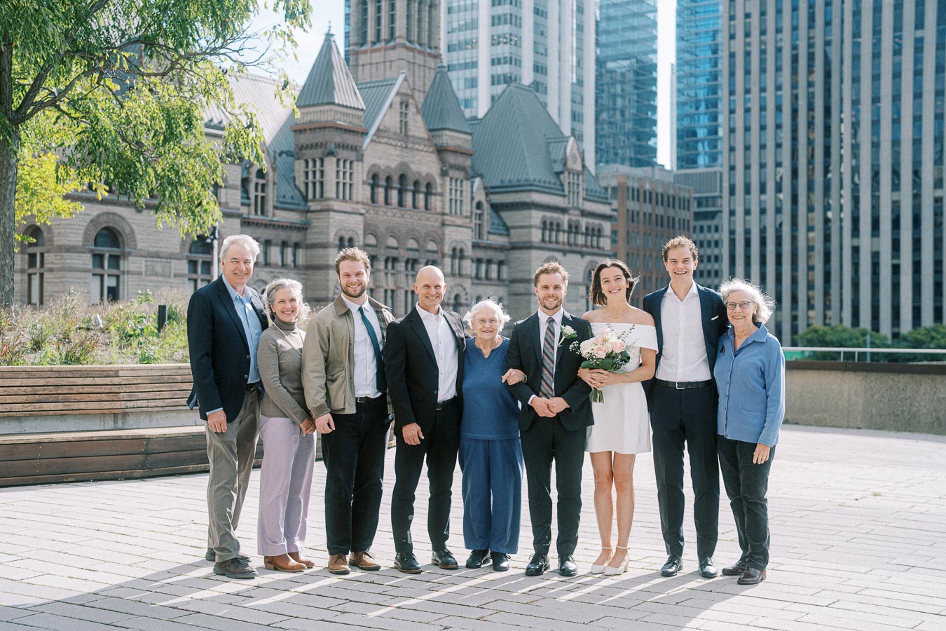A group of people, including a couple dressed in wedding attire, pose together in front of a historic building in a cityscape. The scene features a mix of greenery and tall buildings under a clear sky. Ideal for content about urban weddings, city architecture, or family gatherings.