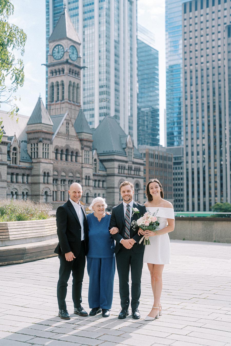 A group of four people posing in formal attire in front of a historic building with a clock tower and surrounded by modern skyscrapers. One person holds a bouquet of flowers. The setting appears to be an urban outdoor plaza.