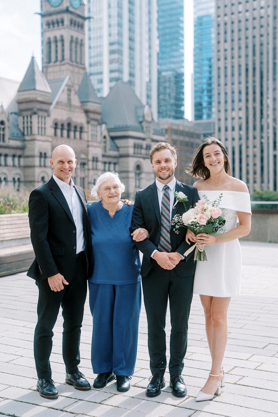 A group of four people posing for a photo in front of historic Toronto architecture, featuring a bride holding a bouquet, a groom in a suit, an elderly woman in blue, and a man in formal wear.