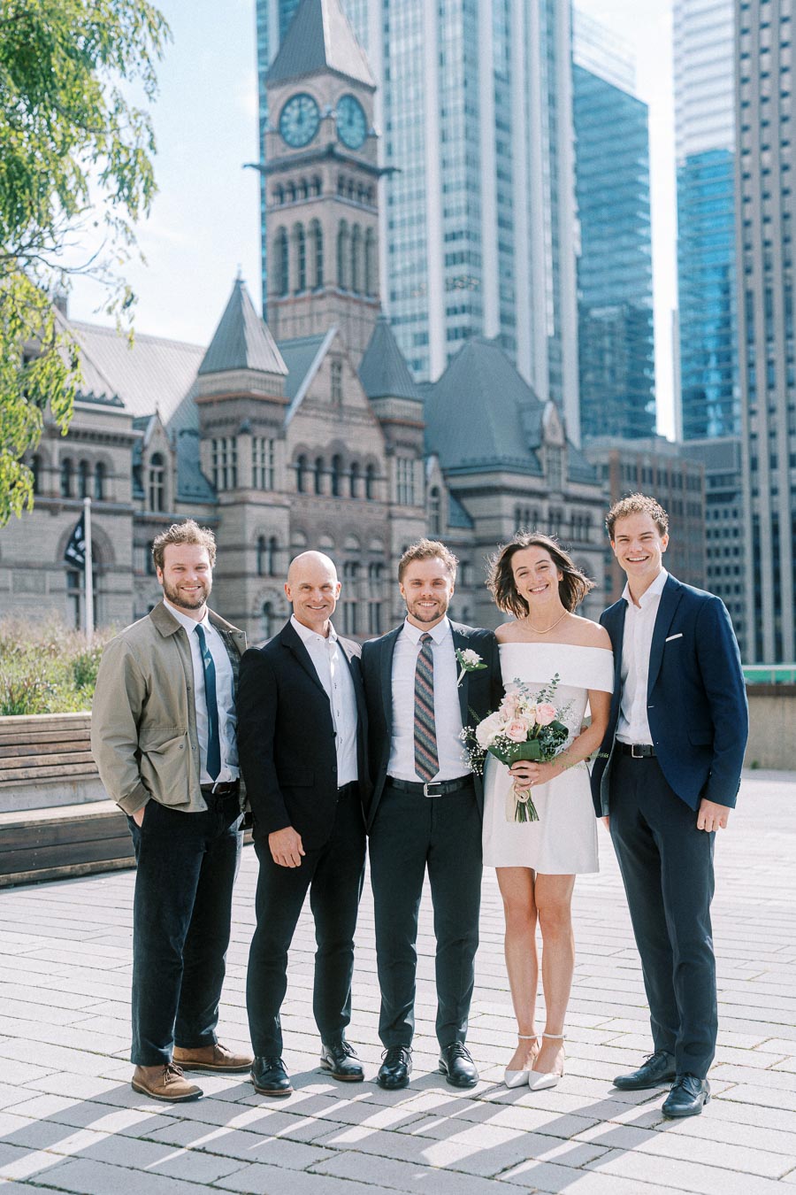 A group of people posing outdoors near a historic building, with one woman holding a bouquet of flowers, dressed in semi-formal attire, in a cityscape setting featuring architectural details.