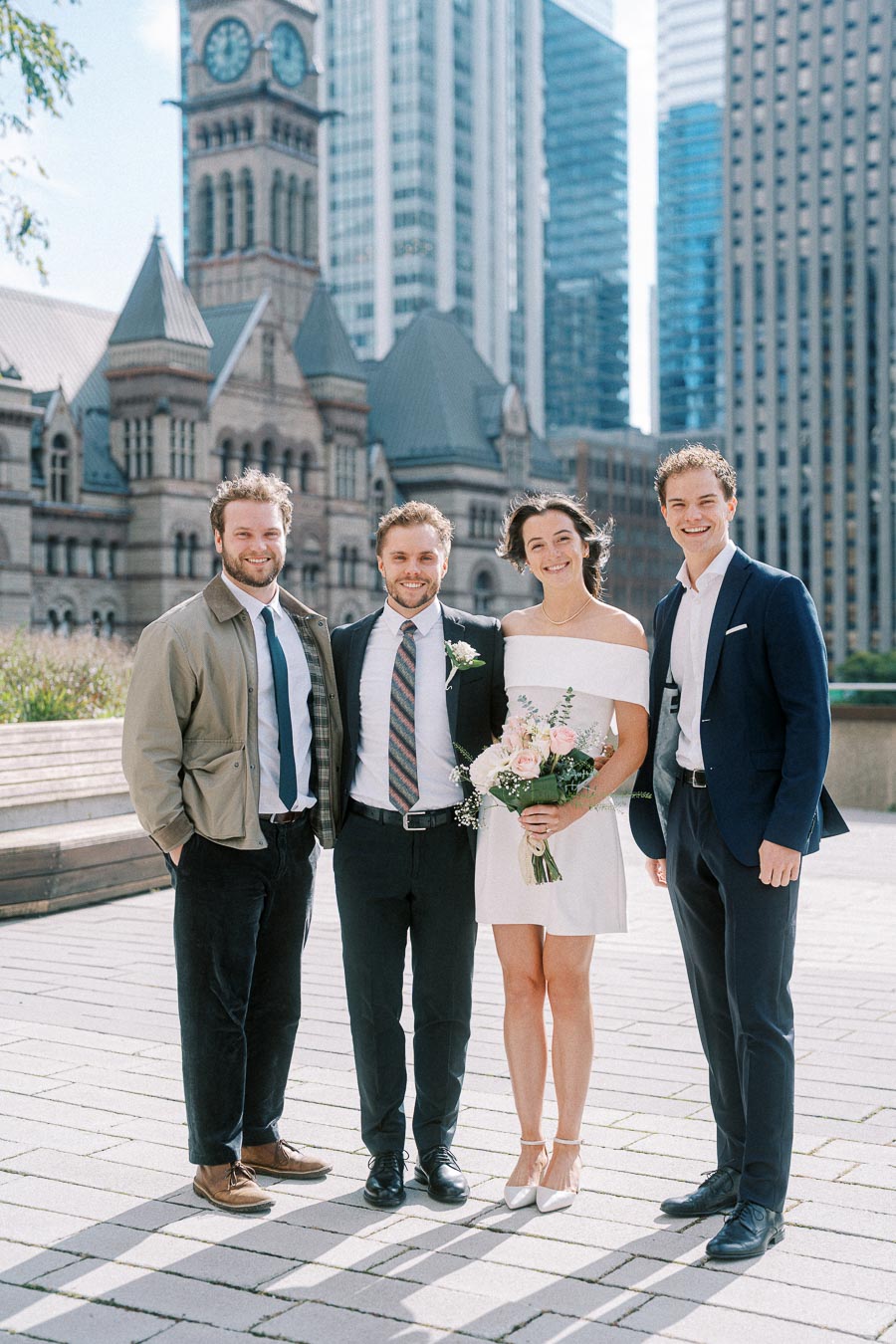 A bridal party poses outdoors in a cityscape setting with historic and modern buildings. The bride holds a bouquet, and three men in suits stand beside her, all smiling.