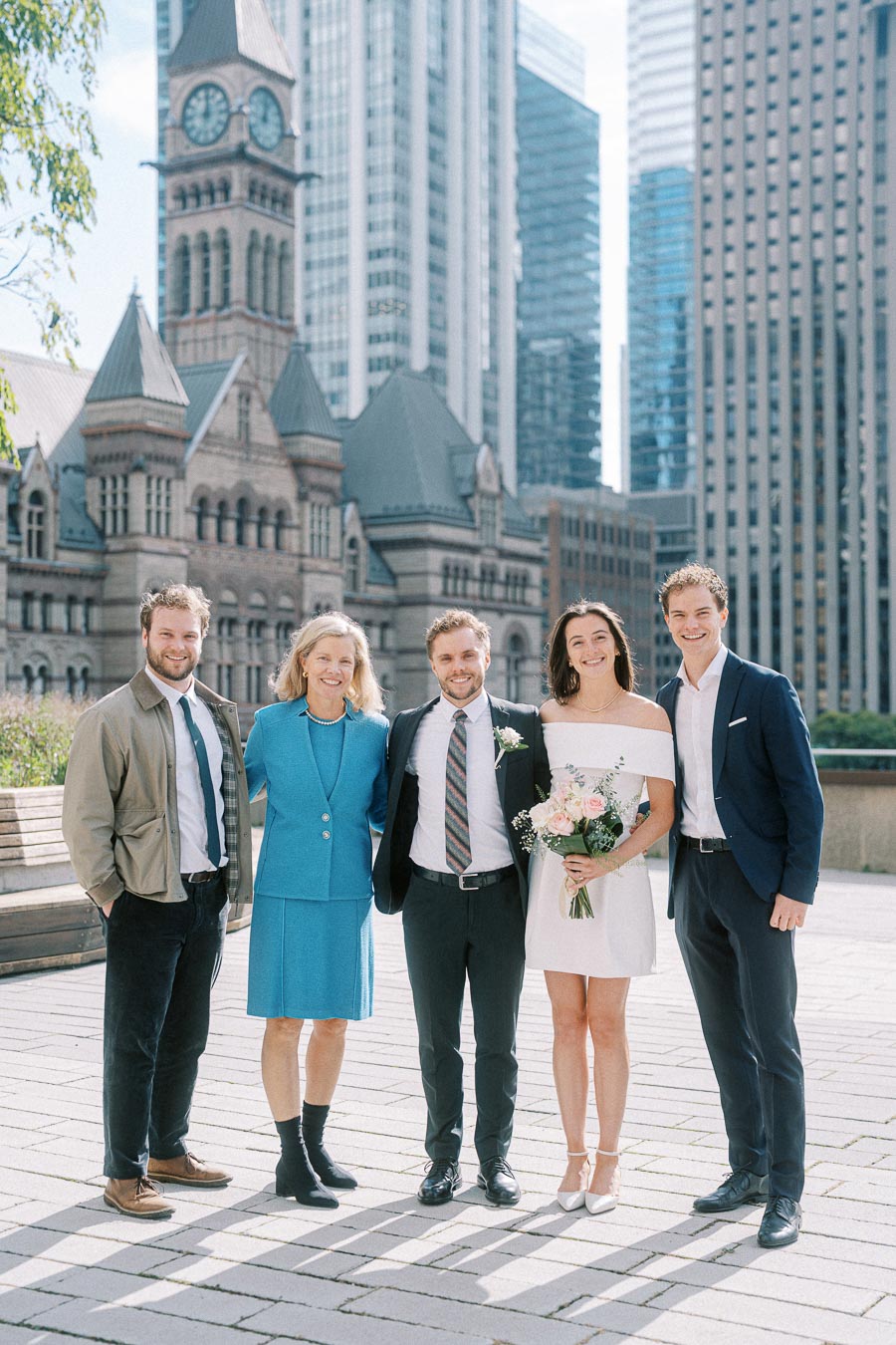 A group of five people dressed formally, with a woman holding a bouquet, standing in front of a historic building and modern skyscrapers in an urban setting.