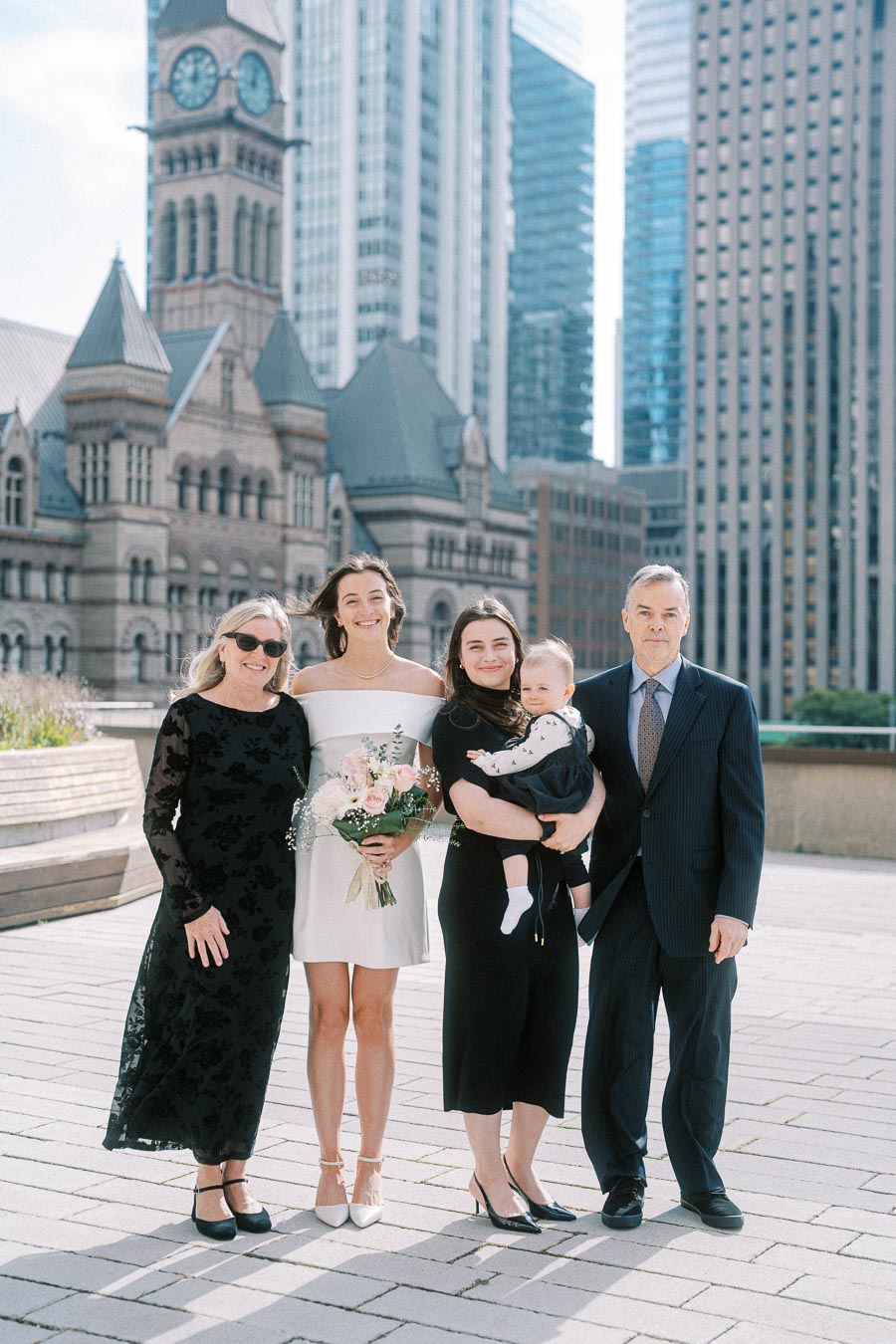 A group of people poses in elegant attire in an urban setting with historic architecture and tall skyscrapers in the background, suggesting a special occasion or celebration.