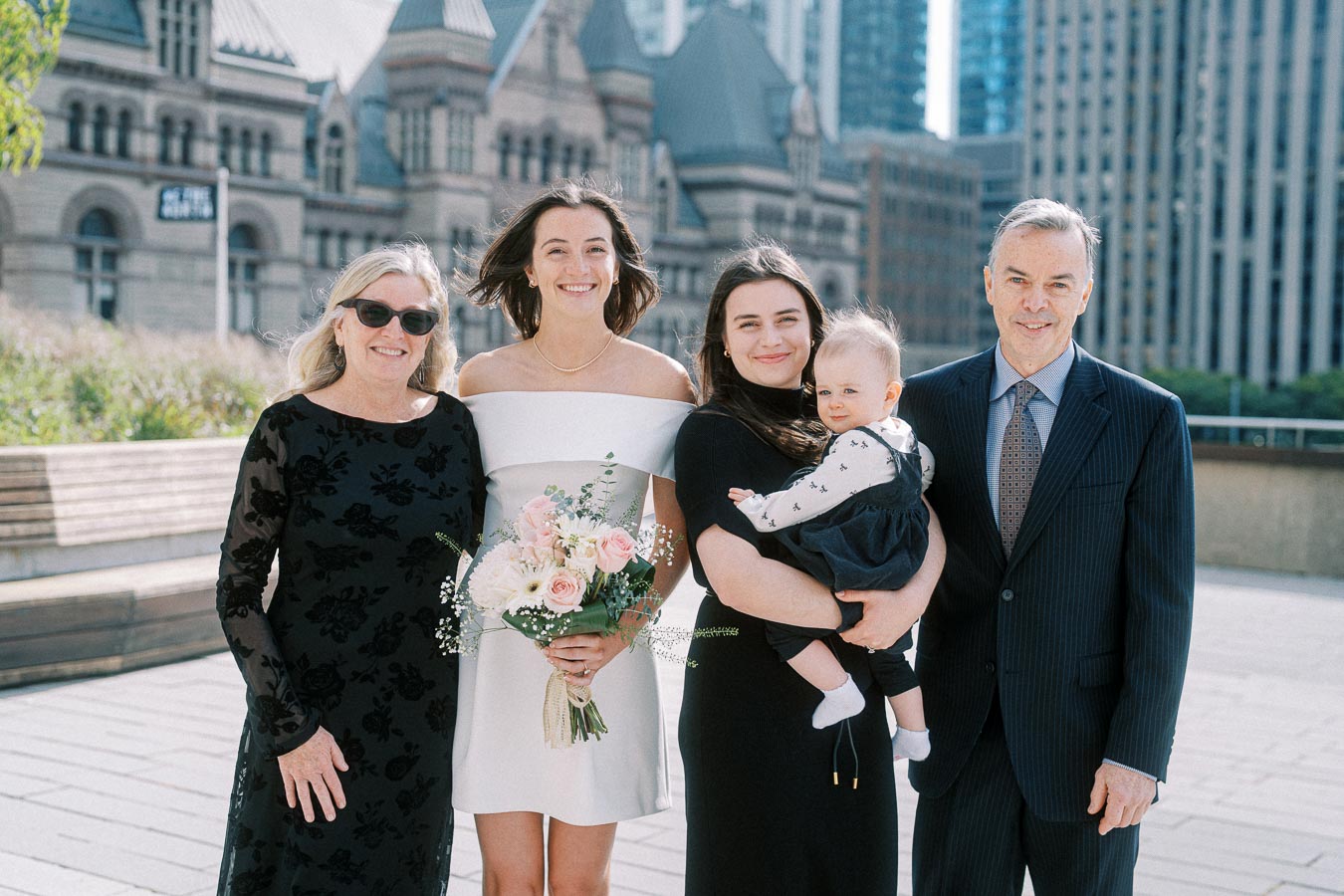 A family gathers for a formal outdoor portrait in an urban setting, featuring a historic building background. The group includes an elegantly dressed woman holding a flower bouquet, a smiling woman holding a baby, flanked by an older couple in formal attire.