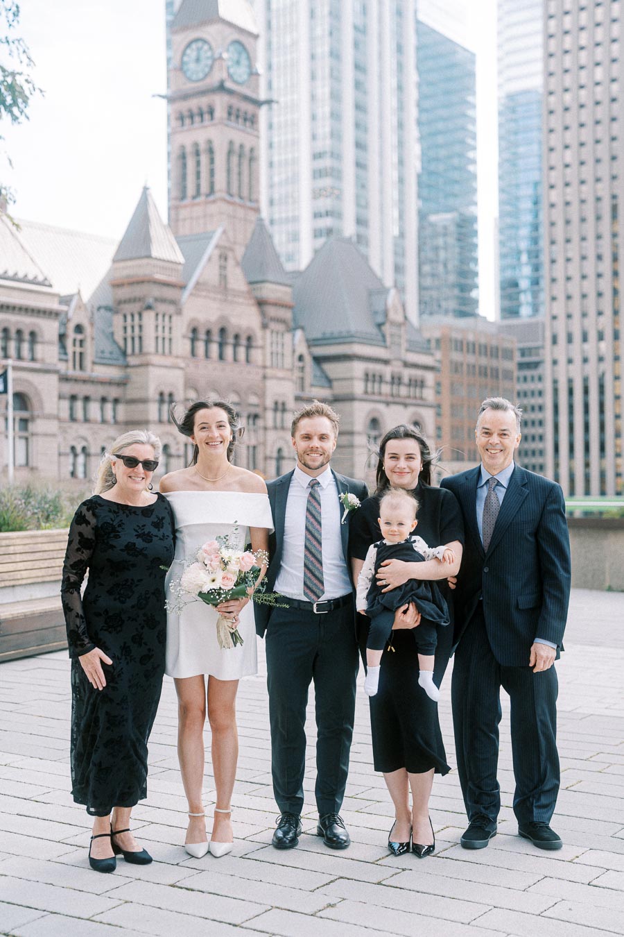 Group photo of elegantly dressed family and friends at an outdoor gathering, featuring a historic clock tower and skyscrapers in the background.