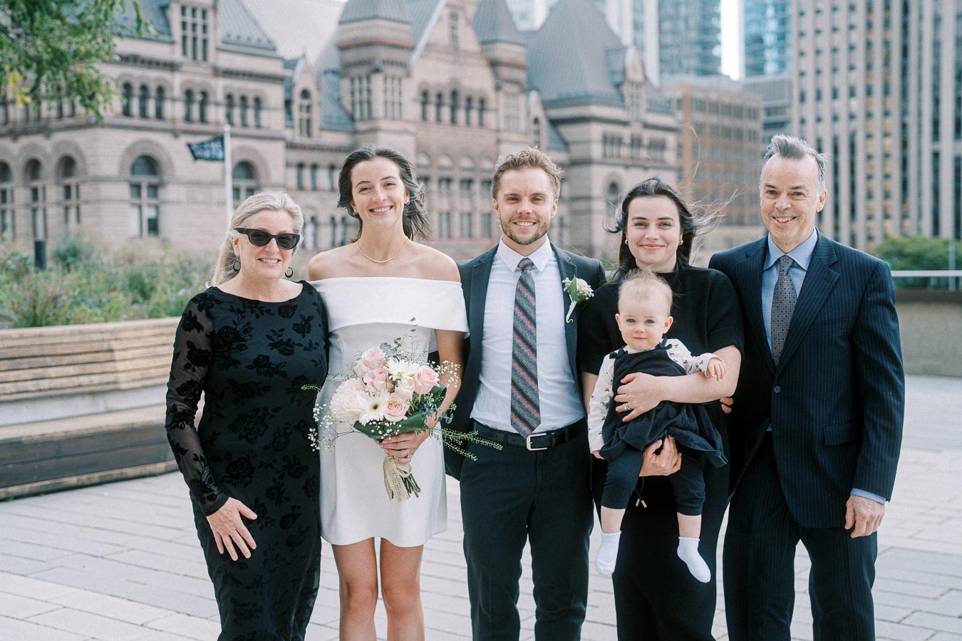 Family posing for a wedding photo, smiling in formal attire, with a historic building in the background.