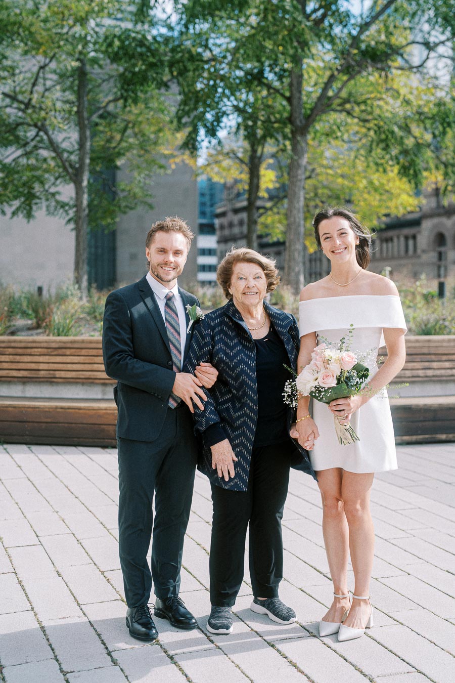 Happy wedding day outdoor portrait featuring a smiling bride in an elegant white dress holding a bouquet, a groom in a formal suit, and an elderly woman, likely a family member, standing together against a backdrop of trees and modern architecture.