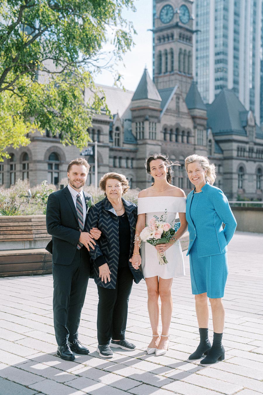 A group photo of four people posing joyfully in front of a historic building with a clock tower. The group includes a person in a white dress holding a bouquet of flowers, accompanied by two individuals in formal attire and one in a blue outfit. The scene is set outdoors on a sunny day with trees and urban architecture as a backdrop.