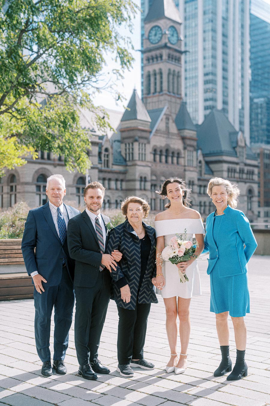 A group of five people, including a bride holding a bouquet, pose together outdoors in front of a historic clock tower and tall buildings on a sunny day.