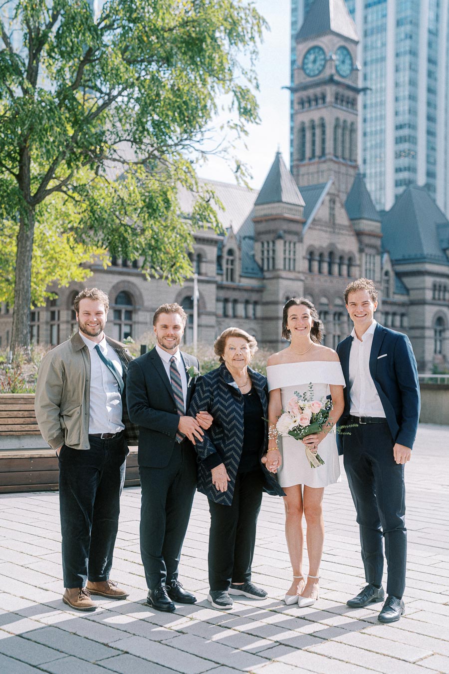 Wedding group portrait outside historical building with clock tower; bride in white dress holding bouquet, surrounded by family members in formal attire, amidst urban backdrop with greenery and architecture.