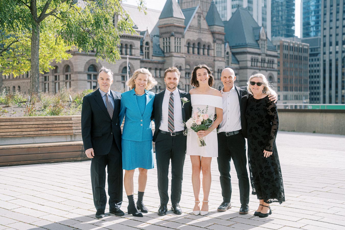 A group of six people, including a bride holding a bouquet, posing outdoors in formal attire with an architectural building in the background on a sunny day.