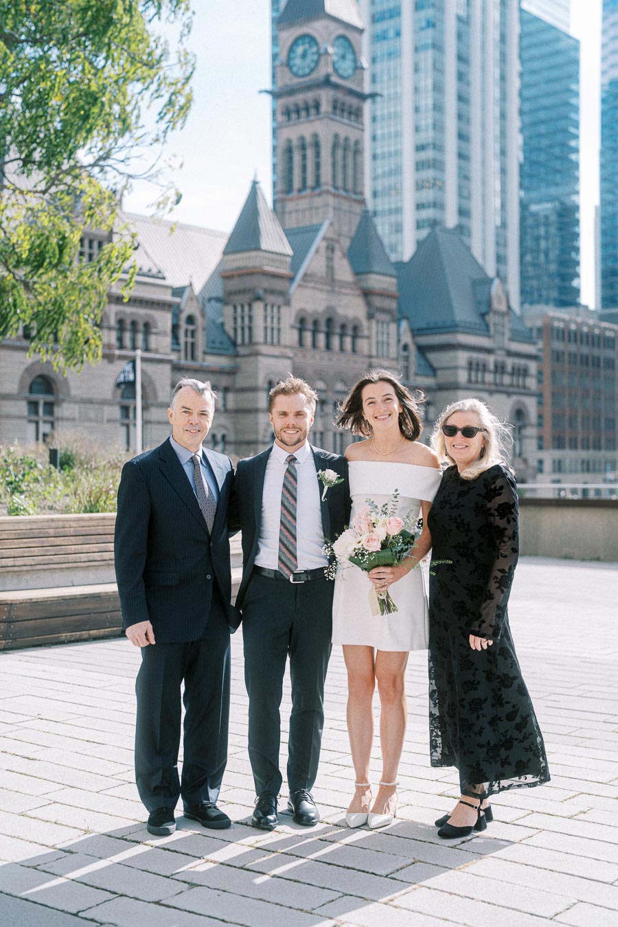 A happy couple posing with two family members in formal attire, standing outdoors in front of a historic building with a clock tower and city skyscrapers in the background. The woman is holding a bouquet of flowers, and the group is smiling on a sunny day.
