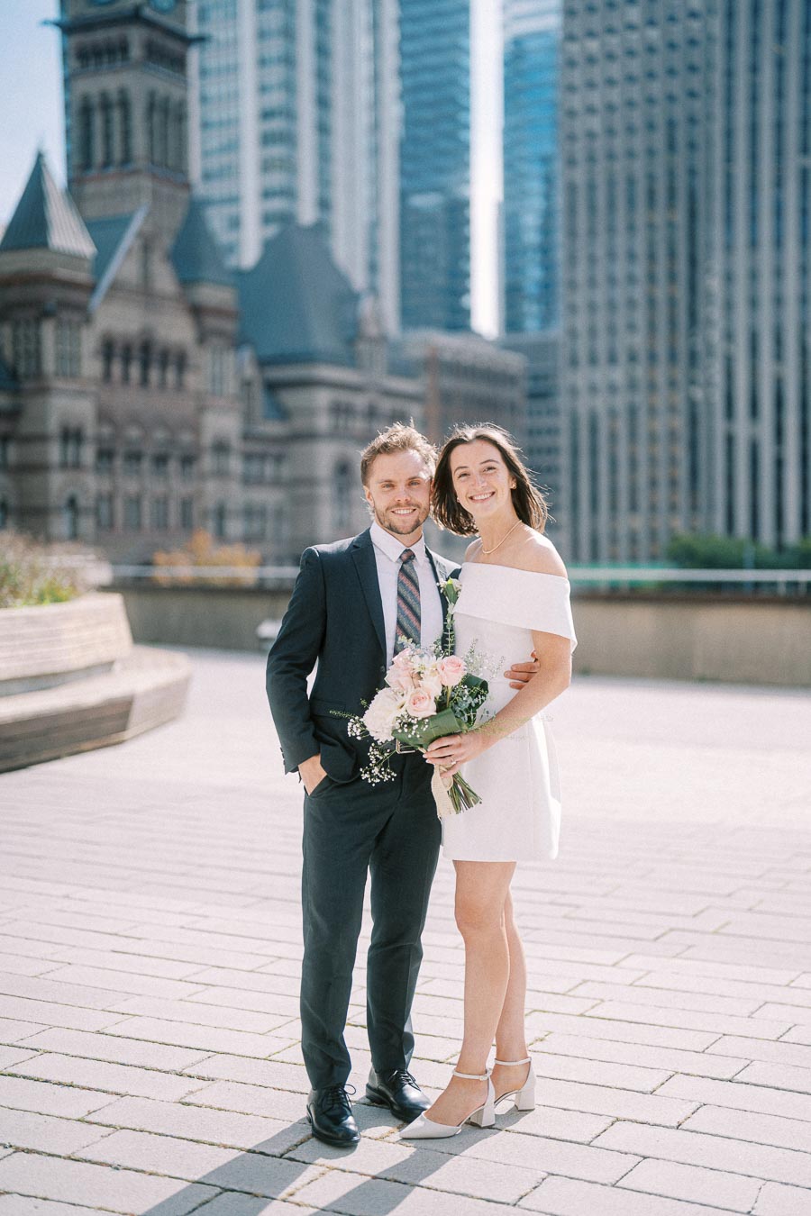 A couple poses for a photo on their wedding day in an urban setting with tall buildings and historic architecture in the background. The bride holds a bouquet of pink and white flowers.