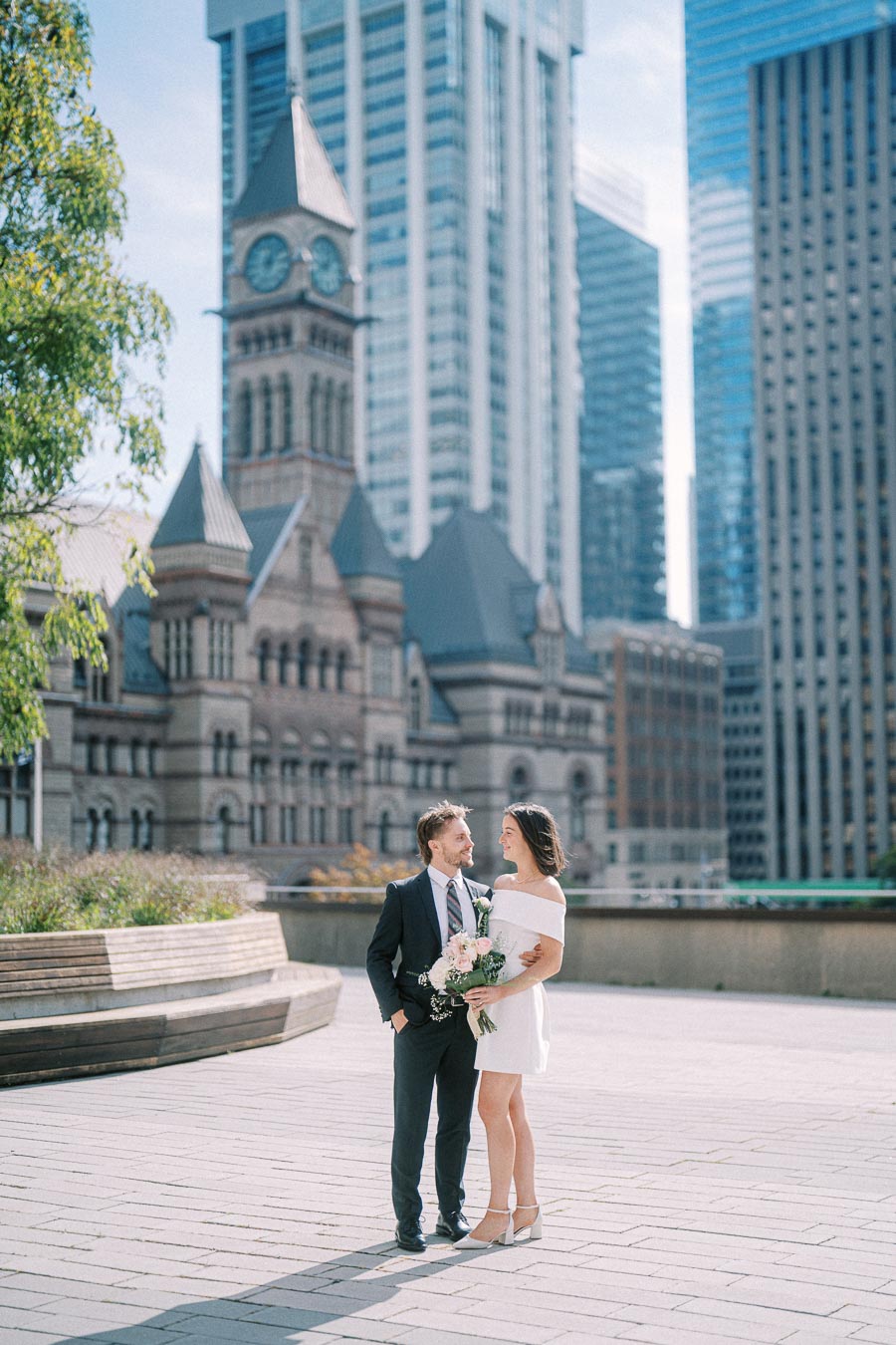 A couple in wedding attire smiles at each other while standing in front of a historic clock tower and modern skyscrapers on a sunny day. The woman is holding a bouquet of flowers, creating a romantic urban setting.
