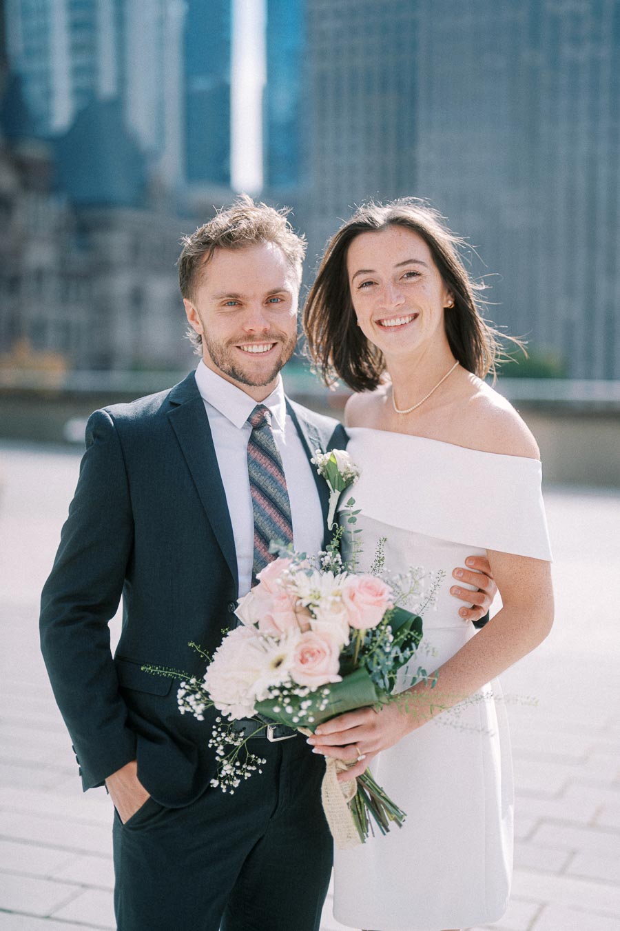 Smiling couple on wedding day in cityscape background, groom in suit and bride holding bouquet of pink and white flowers, enhancing urban wedding theme.