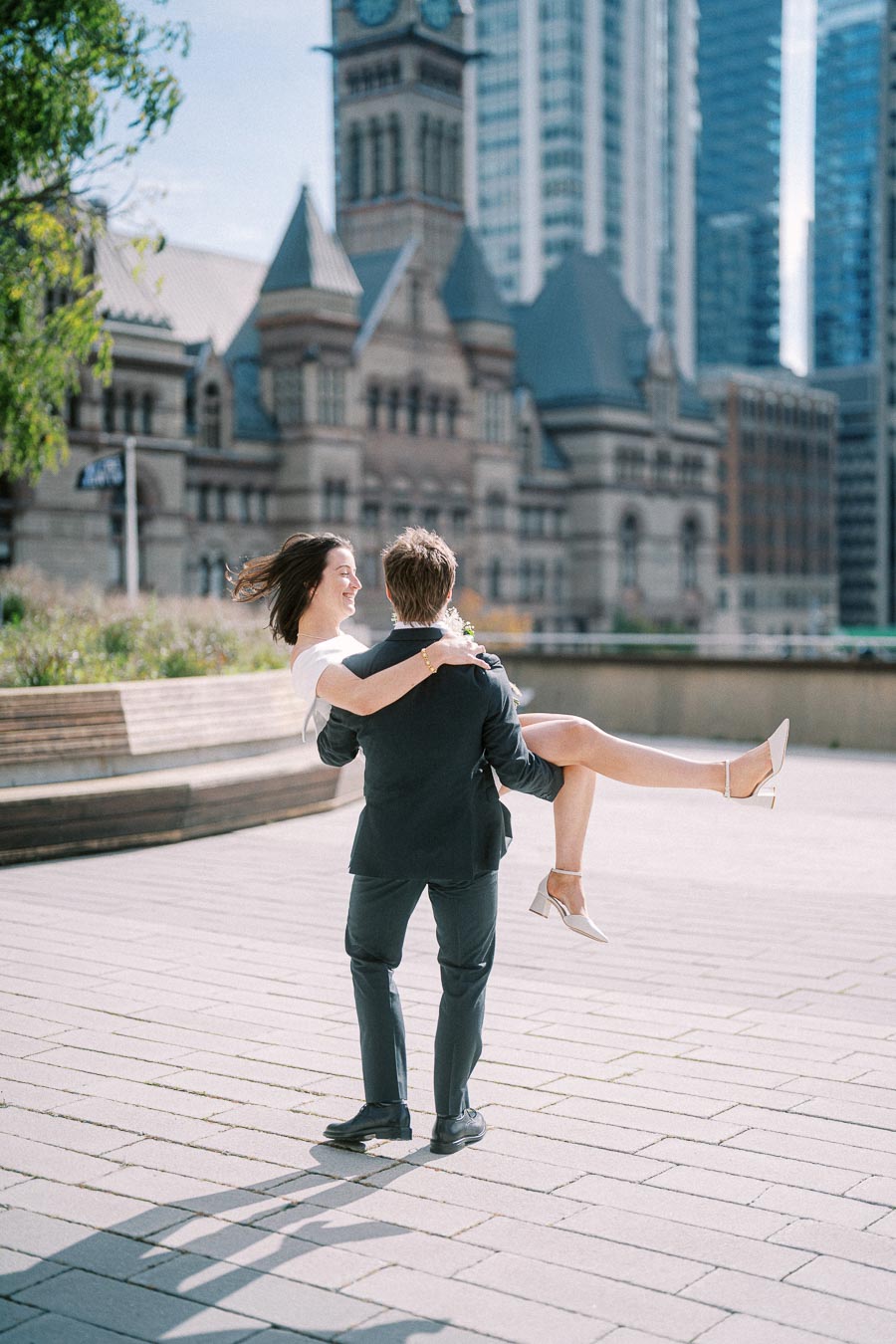 A man in a suit playfully lifts a woman in a white dress against a backdrop of historic architecture and modern skyscrapers in a city plaza.
