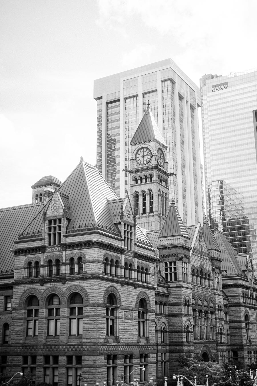 Black and white image of a historic stone building with a clock tower, set against modern skyscrapers in an urban cityscape.