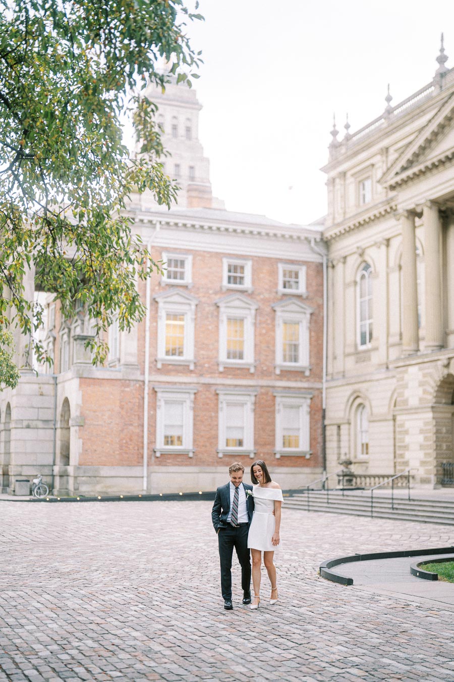 A couple elegantly dressed in formal attire walking in a cobblestone courtyard, surrounded by historic architecture, during a bright, serene day.