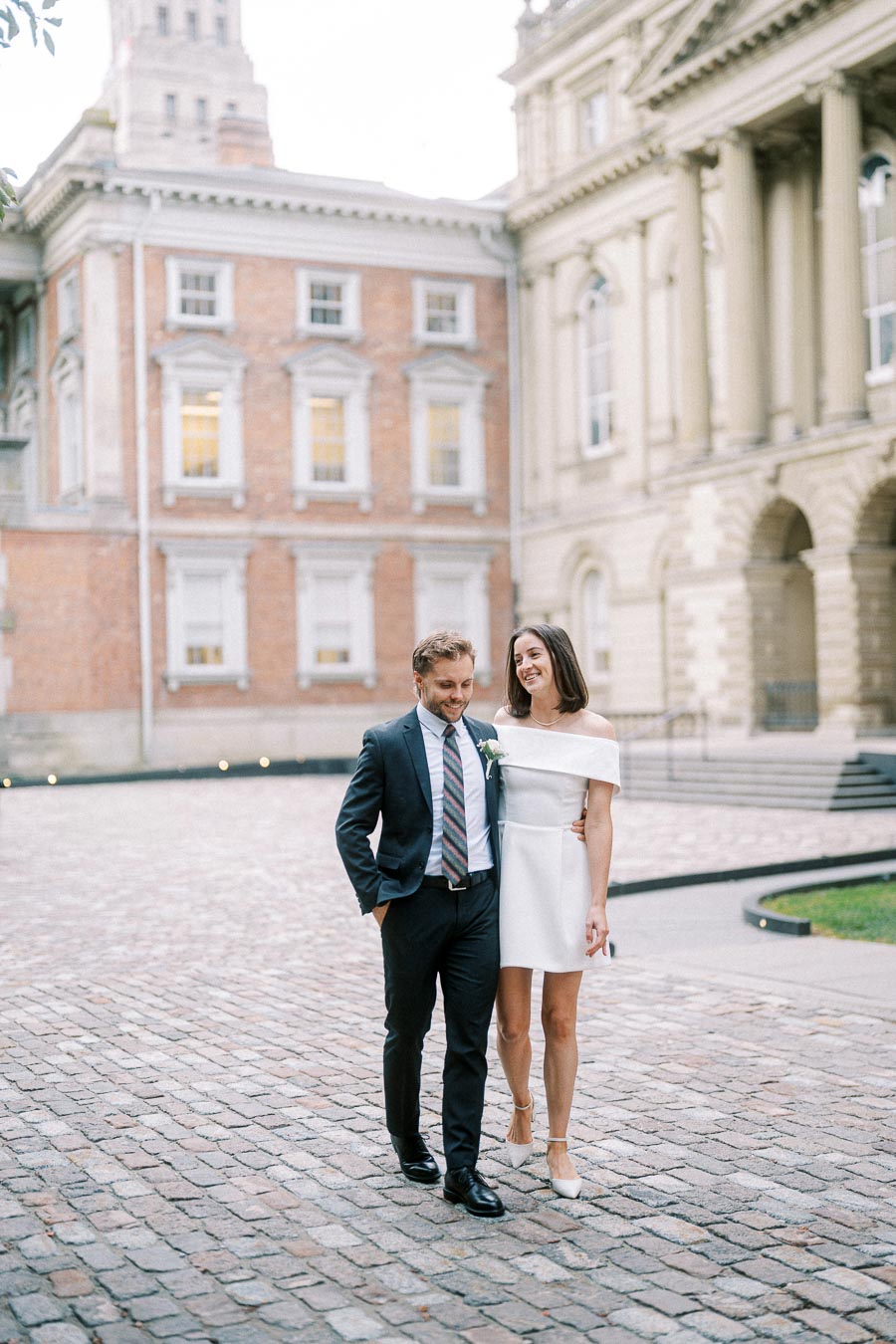 A happy couple, dressed elegantly in formal attire, walks hand-in-hand on a cobblestone pathway between historic buildings, capturing a candid and romantic moment.