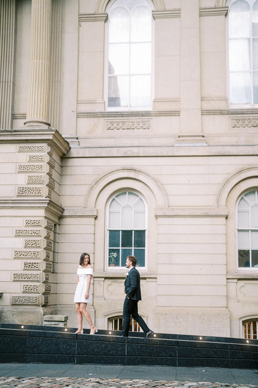 A couple walking on a path in front of an elegant historic building with large arched windows and decorative columns. The woman is in a white dress, the man in a dark suit, both seemingly enjoying a lighthearted moment in an urban setting.