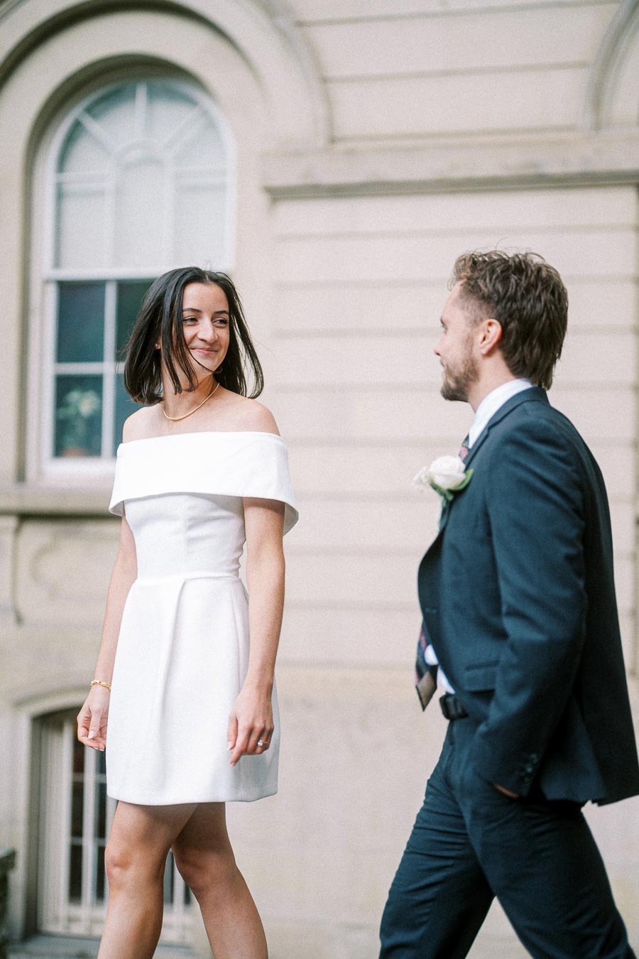 A joyful couple smiling while walking outside a historic building, the woman wearing a white off-shoulder dress and the man dressed in a formal suit.