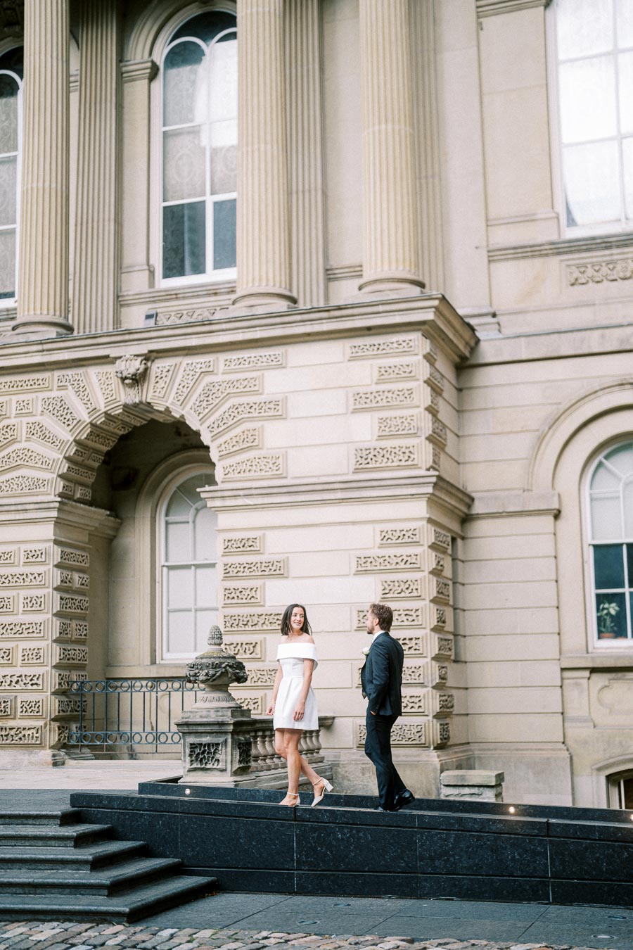 A couple walking on stone steps in front of a historic building with ornate architecture and large arched windows. The woman wears a white dress, and the man is in a suit, engaged in conversation.