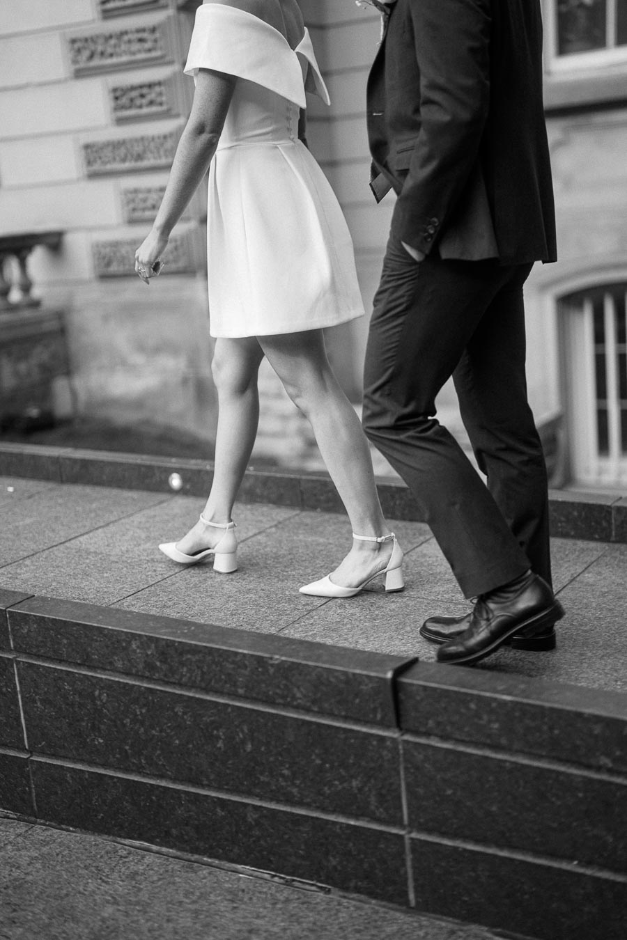 Black and white photo of a couple walking, with the woman in an off-shoulder white dress and heeled shoes, and the man in a dark suit and dress shoes, on a city sidewalk.