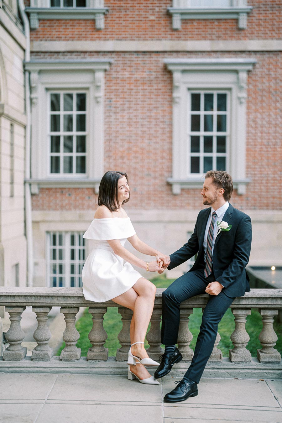 A couple sitting on a stone railing, holding hands and smiling, with a background of a brick building and large windows, dressed in elegant attire for an engagement or wedding photoshoot.