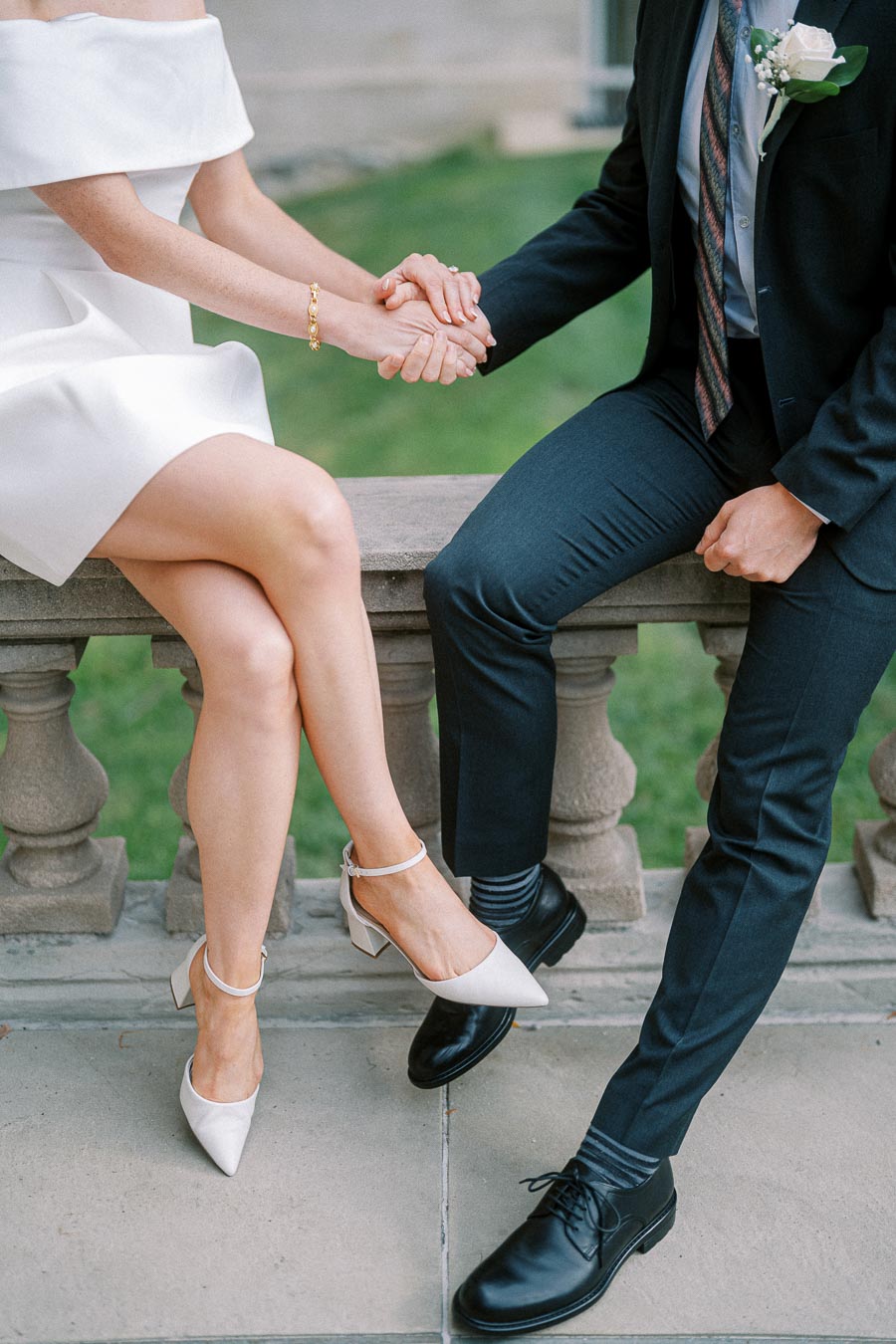 A couple sitting on a stone bench holding hands; a woman in an elegant white dress and white heels, alongside a man in a dark suit and black shoes, showcasing stylish wedding attire and romance.