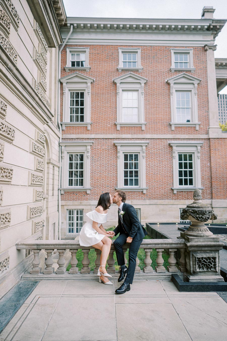 A couple kissing while sitting on a stone railing in front of an elegant, historic brick building, showcasing romantic architecture and a charming outdoor setting.