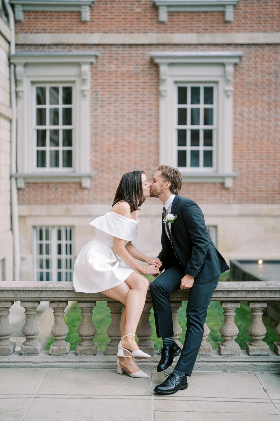 Couple sharing a romantic kiss at a wedding, with bride in white off-shoulder dress and groom in a dark suit, sitting on a stone railing in front of a historic brick building