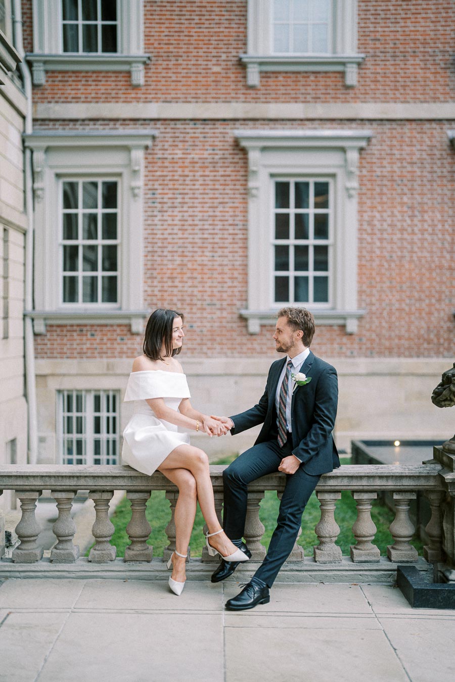 A couple sitting on a stone balustrade in an elegant outdoor setting, with a brick building in the background. The woman is wearing a white dress and the man is in a dark suit, both smiling and holding hands.