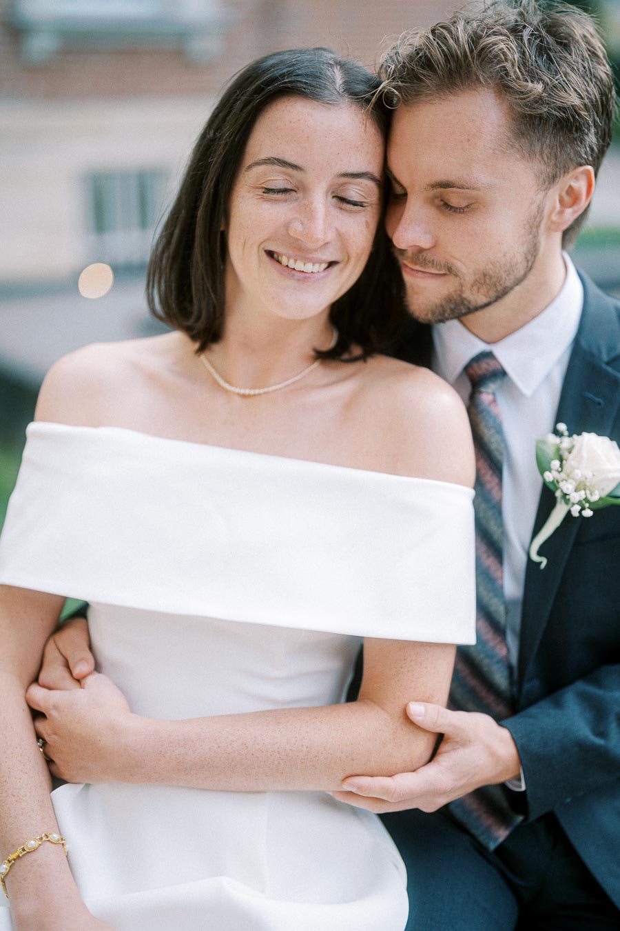 A happy bride in an off-shoulder white wedding gown smiling as she embraces her groom in a navy suit with a rose boutonniere, capturing a tender moment on their special day.